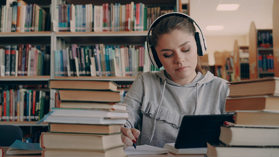 young smiling woman in headphones is sitting at table in university library holding digital tablet and listening to music and writing lecture in notebook