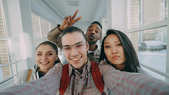 Point of view of four positive cheerful attractive multi-ethnic friends talking selfie photos holding smartphone and having fun laughing while standing in corridor of university.