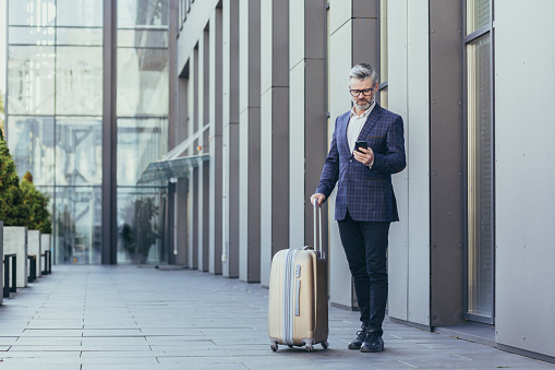 Business trip, senior gray-haired businessman outside hotel using phone, mature boss with big suitcase, investor in business suit and glasses.