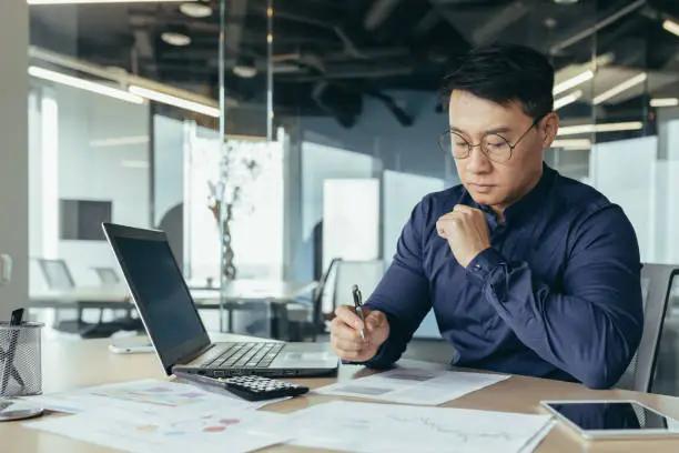 Photo of Thinking asian financier in glasses accountant working with documents and accounts, using laptop, male businessman doing paperwork in sitting at desk inside office building