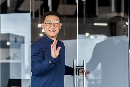 Successful asian worker smiling and looking at camera, man opens door walks into middle of meeting room, holds hand up greets colleagues.