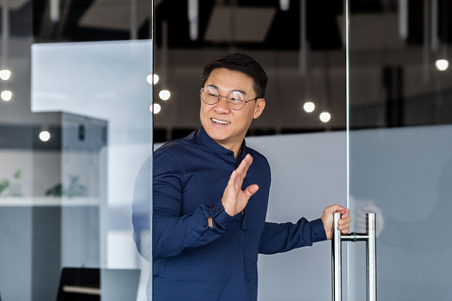 Man walks into meeting room, smiles and greets, Asian boss in glasses and shirt, businessman investor works inside modern office building
