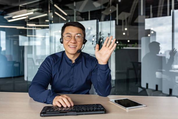 istock Webcam view, portrait of successful tech support and customer service worker, man smiling and looking at camera waving greeting hello, asian working inside modern office building 1434630007