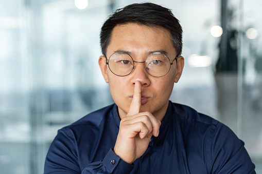 man in glasses and casual shirt businessman looking at camera working inside modern office building asian investor holding finger near mouth demanding silence.