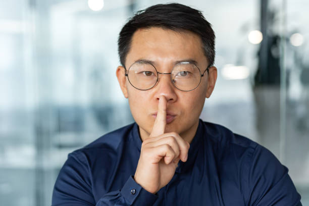 man in glasses and casual shirt businessman looking at camera working inside modern office building asian investor holding finger near mouth demanding silence. - finger on lips imagens e fotografias de stock