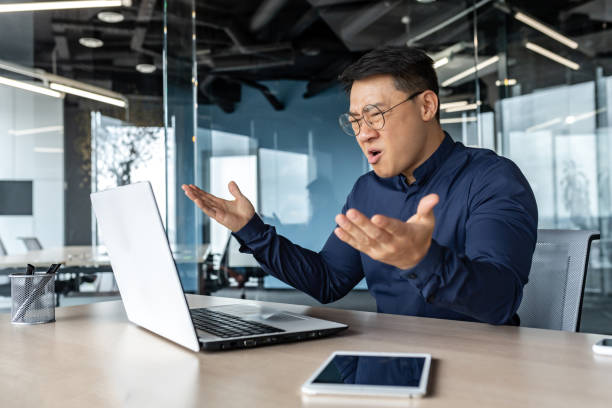 istock Frustrated and angry asian boss yelling at laptop, failure man working inside twisted office building at desk 1434630018
