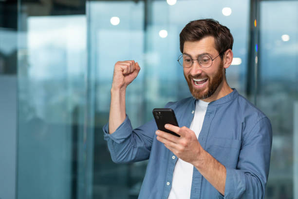istock Successful businessman working in modern office building, man looking at smartphone and reading news celebrating happy news victory businessman in shirt and glasses holding hand up triumph gesture 1434635975