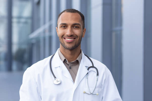 retrato de un exitoso y sonriente médico afroamericano, hombre con bata médica con estetoscopio mirando a la cámara y sonriendo, fuera del hospital moderno - turkish ethnicity white black contemporary fotografías e imágenes de stock