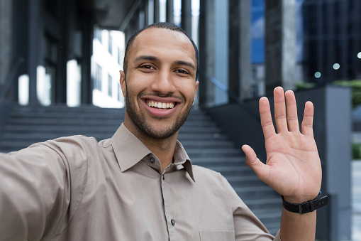 Young african american man looking into smartphone camera, talking on video call using app on phone, smiling and happy waving hand gesture of greeting, businessman outside modern office building.