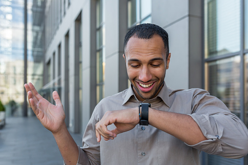 Young successful arab businessman talking on smart watch and smiling, app developer programmer walking outside modern office building, worker in casual shirt.