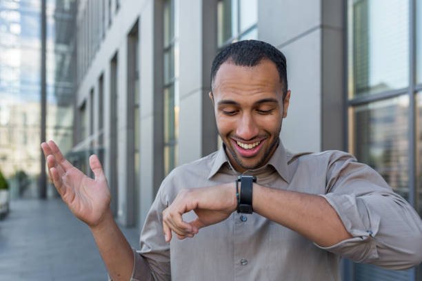 jovem empresário árabe de sucesso falando em relógio inteligente e sorrindo, programador desenvolvedor de aplicativos andando fora do moderno edifício de escritórios, trabalhador de camisa casual - mid adult men smart casual white happiness - fotografias e filmes do acervo