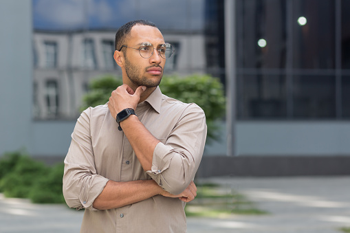 Young pensive businessman in glasses outside modern office building, man thinking and looking away, office worker in shirt.