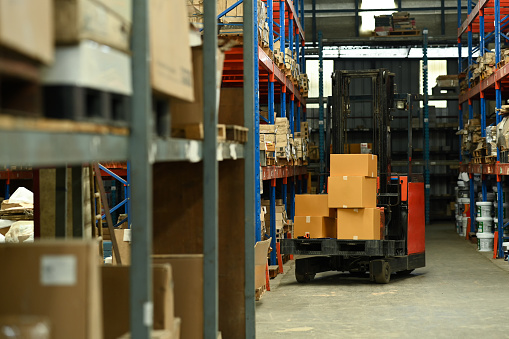 Forklift truck parking in large logistic distribution warehouse full of shelves with cardboard boxes.