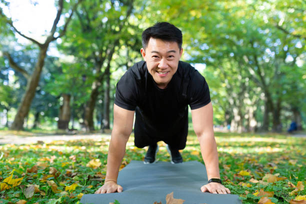 retrato de atleta masculino feliz, asiático mirando a la cámara y sonriendo haciendo ejercicios físicos, flexiones y plancha en el parque de otoño en un día soleado - filipino ethnicity asian ethnicity men male fotografías e imágenes de stock