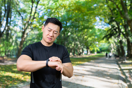 unhappy and upset male athlete looking at fitness with smartwatch, bad workout result, asian man exercising in autumn park on day.