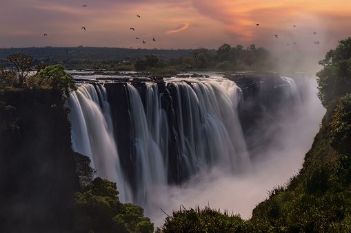 An aerial view of Mosi-Oa-Tunya Falls waterfall in Zambia during sunset with birds flying around