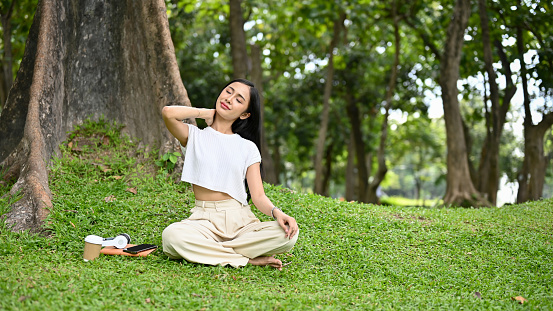 Relaxed and happy young Asian female in casual clothes sitting under the tree, stretching and massaging her neck, getting fresh air in the park. Wellbeing lifestyle