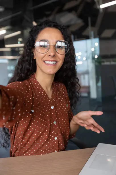 Vertical shot, video call from office hispanic business woman working inside modern office building, talking remotely with friends using app on phone for video call