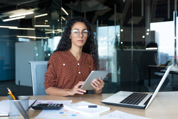 istock Thinking businesswoman with tablet computer looking seriously at camera, Hispanic woman working inside modern office building with documents, 1434661762