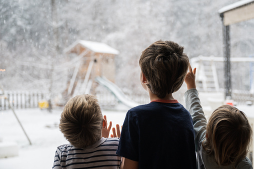View from behind of three children, siblings, looking out the window into a beautiful winter nature with snow falling down the sky.
