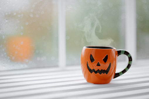 Halloween celebration concept with spooky jack o lantern big cup at autumn season. All hallows eve party symbol hot beverage in orange pumpkin mug on wooden windowsill at window background