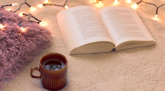Concept of relaxation and hygge on a winter day. A cup of black, hot coffee and an open book on the bed with Christmas lights. Copy space.