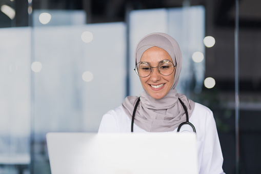 Close-up photo portrait of a female doctor in a hijab, a Muslim woman works in the office of a modern clinic, a female doctor in a gown and glasses uses a laptop.