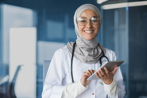 młoda piękna muzułmańska lekarka w hidżabie uśmiechająca się i patrząca na aparat pracuj�ący z tabletem - healthcare and medicine digital tablet medical exam doctor zdjęcia i obrazy z banku zdjęć