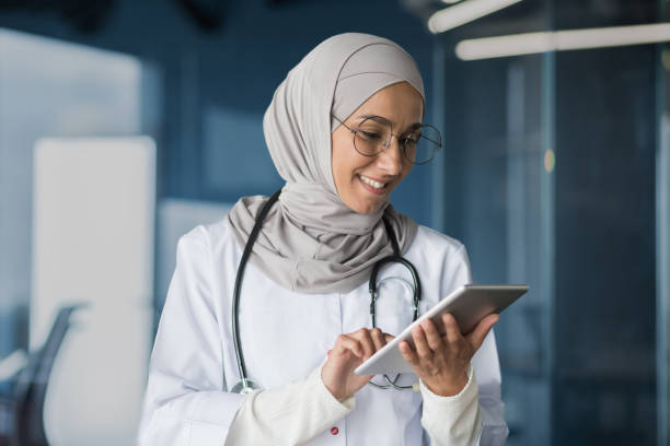 la doctora con hijab trabaja en la oficina de la clínica moderna, la doctora musulmana usa una tableta, una enfermera con bata blanca médica y gafas con estetoscopio - computer medical student hospital nurse fotografías e imágenes de stock