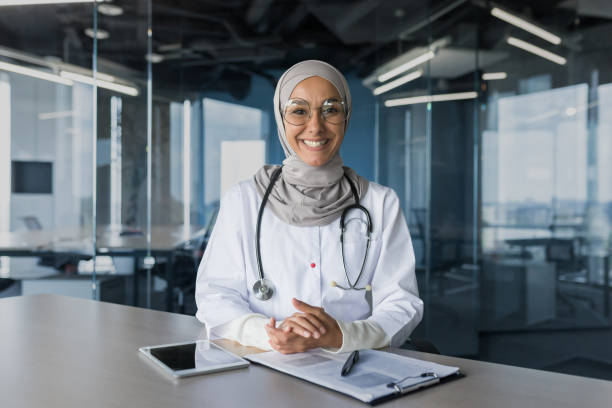 portrait d’une femme médecin musulmane à succès en hijab, femme arabe travaillant dans un bureau de clinique moderne, souriant et regardant la caméra, femme médecin en lunettes et blouse médicale blanche avec stéthoscope - service computer training office photos et images de collection