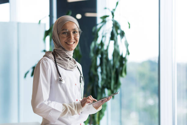 retrato de uma médica muçulmana em um hijab com um comprimido nas mãos, o médico trabalha no consultório de uma clínica moderna, sorri e olha para a câmera - healthcare and medicine digital tablet medical exam doctor - fotografias e filmes do acervo
