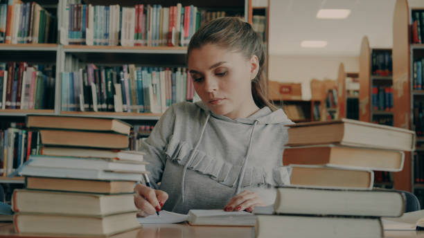 istock Concentrated young female student reading books and writing in notebook during working at school project in university library 1434683633