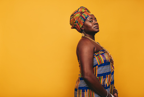 Stylish African woman wearing traditional clothing against a yellow background. Mature black woman dressed in colourful Kente cloth and golden jewellery. Confident woman embracing her rich culture.