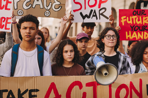 Youth activists holding up banners and placards during an anti-war rally. Group of multicultural young people marching for world peace. Teenage demonstrators protesting against war and violence.
