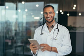 Portrait of happy and successful african american doctor man working inside office clinic holding tablet computer looking at camera and smiling wearing white coat with stethoscope