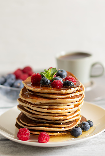 A stack of pancakes on a white plate with raspberries, blueberries and a cup of tea on a light background. Side view, vertical, close-up.