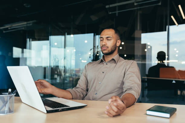 Successful businessman working inside modern office building, man meditating with eyes closed in lotus position sitting on chair Successful businessman working inside modern office building, man meditating with eyes closed in lotus position sitting on chair. managing stress stock pictures, royalty-free photos & images