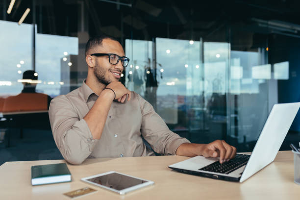 uomo indiano felice e di successo che lavora in un moderno edificio per uffici, programmatore in occhiali che utilizza il laptop per scrivere codice, uomo d'affari in camicia sorridente e felice - business person vanishing point businessman black and white foto e immagini stock