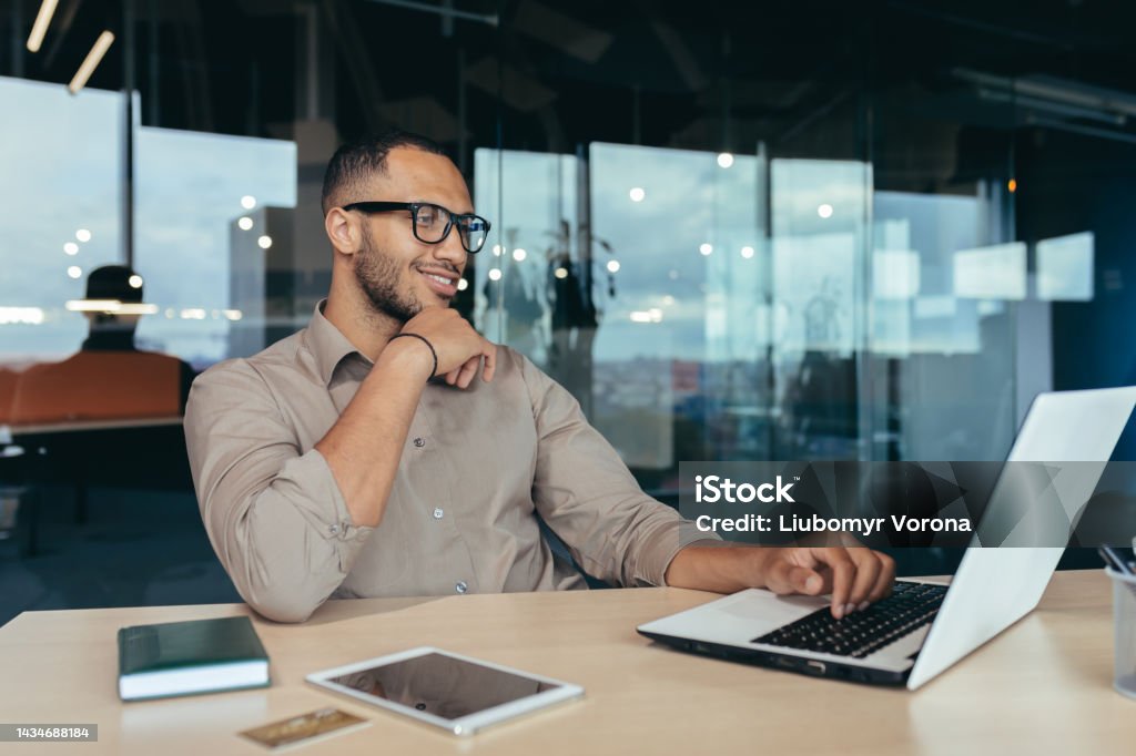 Happy and successful Indian man working in modern office building, programmer in glasses using laptop to write code, businessman in shirt smiling and happy Happy and successful Indian man working in modern office building, programmer in glasses using laptop to write code, businessman in shirt smiling and happy . Men Stock Photo