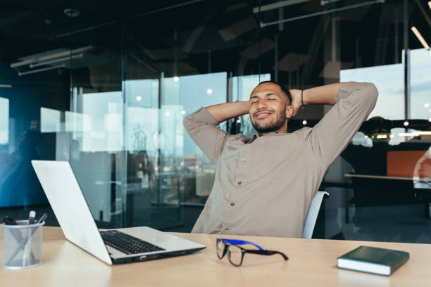 istock mixed race man relaxing in office with hands behind head looking out window and smiling, businessman daydreaming inside modern office building, successful investor happy with work 1434688193