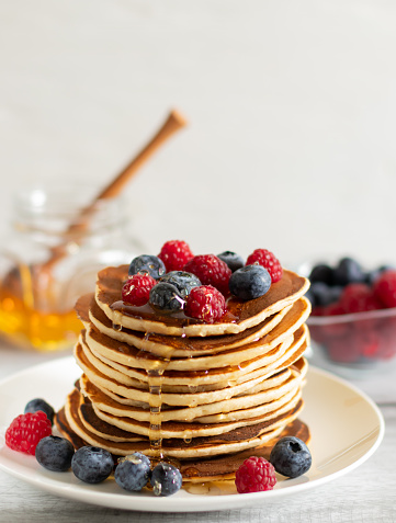 A stack of pancakes on a white plate with raspberries, blueberries and a jar of honey on a light background. Side view, vertical, close-up.