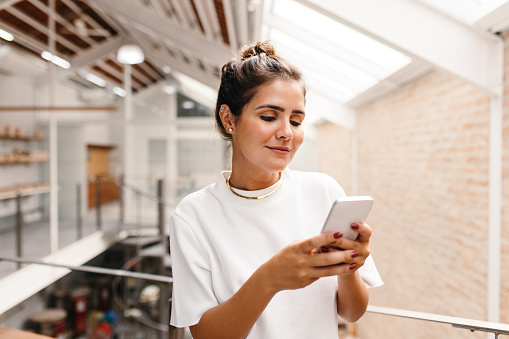 Self-employed businesswoman using a smartphone while standing in a warehouse. Creative female entrepreneur sending a text message. Young woman running an online small business.