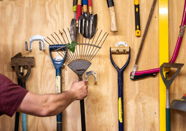 Man's arm taking lawn and leaf rake off wooden wall with various hanging DIY garden tools in shed. Man's arm takes lawn and leaf rake off wooden wall with various hanging DIY garden tools inside shed. Tools include shovel, hammer, fork, trowel, spirit level measure, saw etc. shed stock pictures, royalty-free photos & images