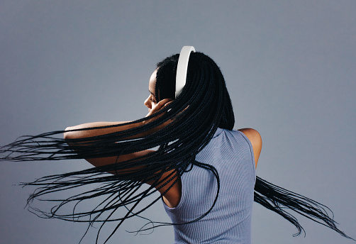 Young woman flipping her braids while listening to music on wireless headphones in a studio. Young woman dancing to an audio playlist while standing against a grey background.