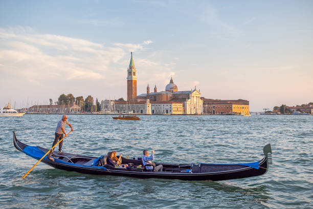 Venice, Italy - October 6th 2022: Traditional dressed gondolier sailing tourists up a broad canal in front of a old church in the center of Venice