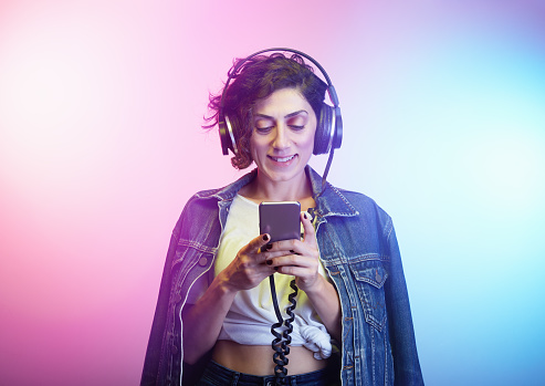 Studio shot of an attractive young woman wearing headphones and denim jacket, searcing and listening to music against a colorful background