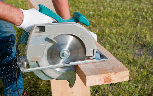 A male joiner works with a hand-held circular saw. A builder is sawing a board at the construction site of a house.