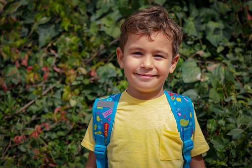 4 years old boy, going to kindergarten. Backpack, yellow t-shirt, sportswear. Boy with cute expression.Background is green,leaves,nature.Model bust, portrait, looking at camera.