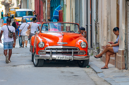 Havana, Cuba - September 16, 2022: People walking down a narrow street. Cars are parked on one side of the street, and a woman sits beside the road in front of a house.