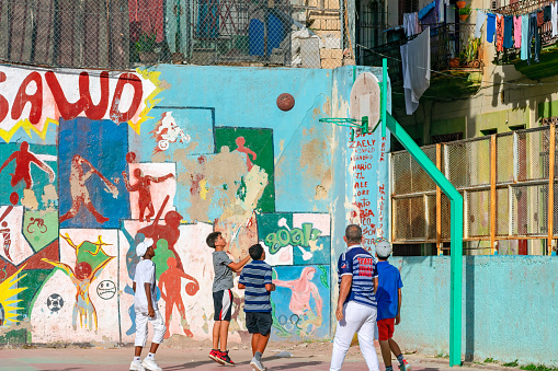 Havana, Cuba - September 16, 2022:A group of boys plays basketball in a playground.  There is graffiti on the walls around the area, and a passerby is observing the game.